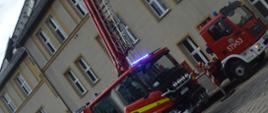 Ćwiczenia w ZOL. Zdjęcie przedstawia 2 wozy strażackie na tle budynku zakładu opiekuńczo-leczniczego. Jeden z nich to ciężki wóz ratowniczo-gaśniczy, ktory ma otwarte dwie tylne skrytki i włączone niebieskie światła błyskowe, a drugi to specjalny pojazd z podnośnikiem hydraulicznym. 