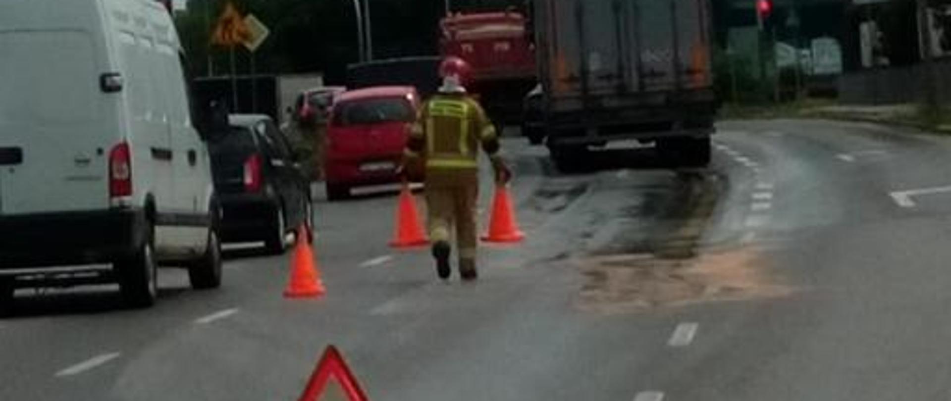 Zdjęcie przedstawia ulicę Łódzką w Kielcach. Lewym pasem ruch odbywa się płynnie, środkowy pas zajęty jest przez powstałą plamę oraz działających przy jej usuwaniu strażaków. Widać plamy sorbentu oraz wydzielenia bezpieczeństwa wykonane za pomocą pachołków i trójkątów ostrzegawczych.