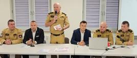 Mężczyzna w mundurze Państwowej Straży Pożarnej przemawia do mikrofonu, stojąc za stołem konferencyjnym. Przy stole siedzą funkcjonariusze PSP oraz dwóch mężczyzn w garniturach. Na stole znajdują się dokumenty, laptop i mikrofony.