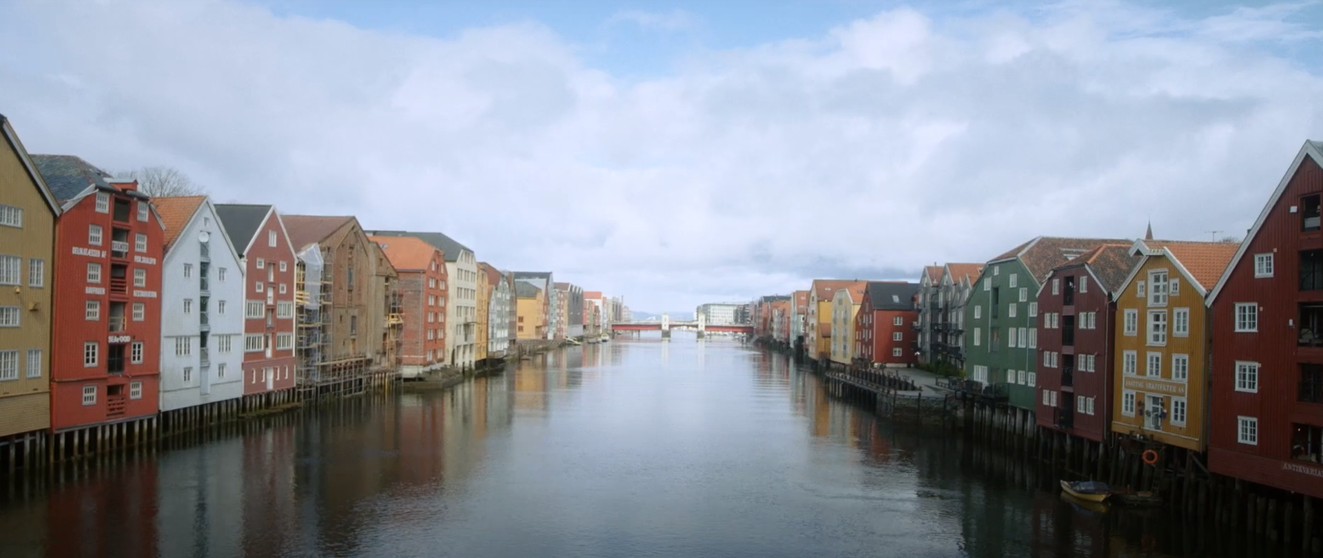 Rzeka na wprost na szerokość zdjęcia. Na brzegach rzeki kolorowe norweskie domki czteropiętrowe. W tle most na rzece. Niebieskie zachmurzone niebo.