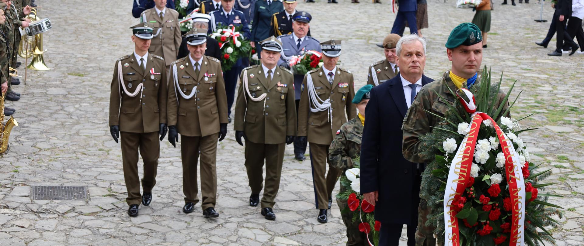 Żołnierz niesie wieniec, za nim mężczyzna i grupa żołnierz.