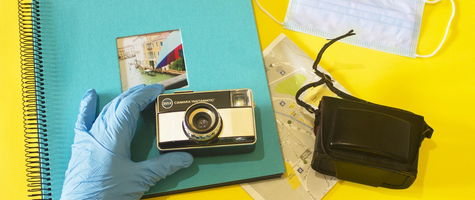 Na żółtym tle leżą: album ze zdjęciami, mapa, maseczka jednorazowa ochronna, etui do aparatu fotograficznego, a ręka w jednorazowej rękawiczce ochronnej trzyma aparat fotograficzny nad albumem do zdjęć 