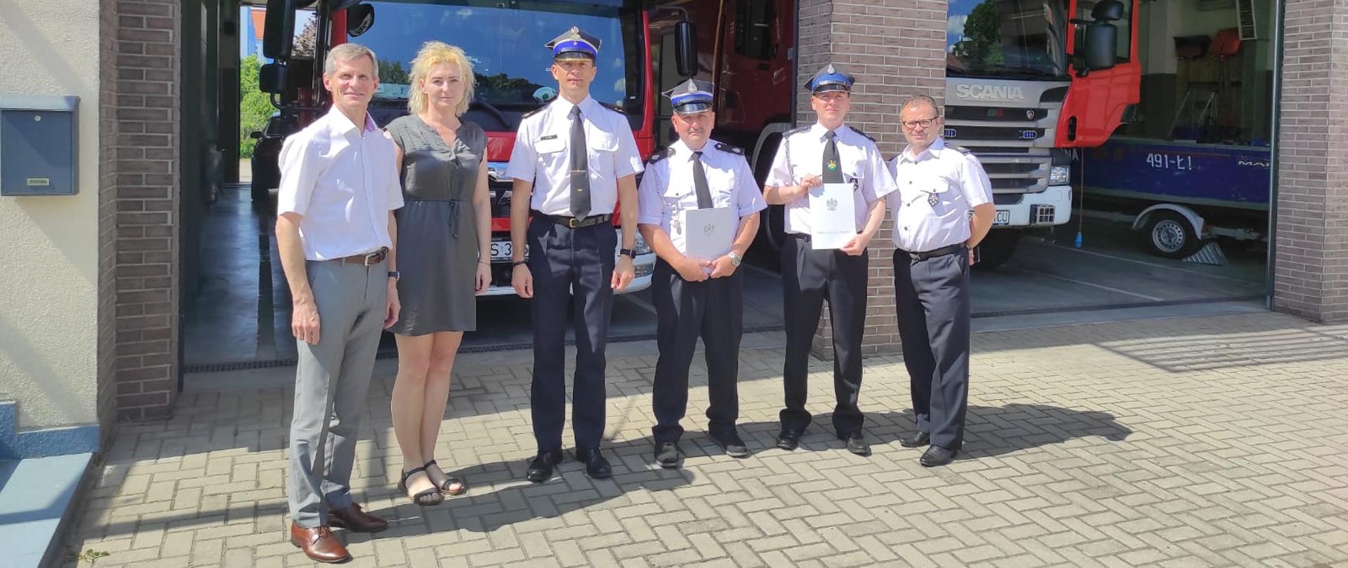 Na zdjęciu 6 osób w tym funkcjonariusze straży pożarnej ustawieni frontem do zdjęcia. Część z funkcjonariuszy trzyma w ręku akty przekazania - teczki. W tle garaże oraz pojazdy pożarnicze.