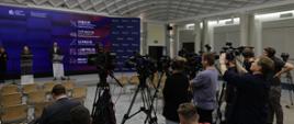 Minister Katarzyna Pełczyńska-Nałęcz and Deputy Minister Jan Szyszko stand on stage in front of microphones. Journalists can be seen standing on the right.