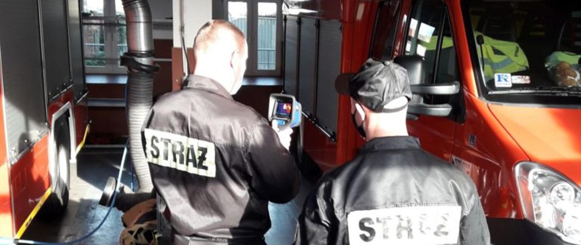 Dwóch strażaków z jednostki ratowniczo gaśniczej w Lubsku ubranych w czarne ubrania koszarowe testuje nową kamerę termowizyjną w garażu jednostki. Na zdjęciu widać dwa czerwone samochody strażackie zaparkowane w garażu.
