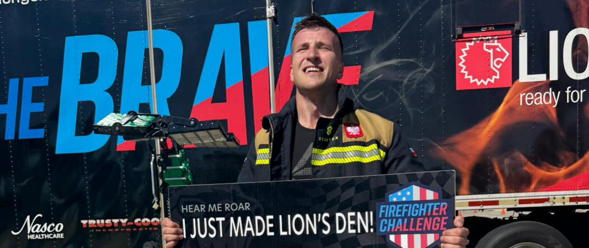 Na zdjęciu widoczny jest strażak w ubraniu specjalnym po zakończonym starcie w zawodach Firefighter Challenge. Uczestnik trzyma pamiątkową tablicę z napisem „I Just Made Lion’s Den!”, która potwierdza ukończenie wymagającej konkurencji. Na ubraniu widoczna jest naszywka z polską flagą, podkreślająca reprezentowanie Polski podczas zawodów. W tle znajduje się charakterystyczna naczepa z logo Firefighter Challenge i partnera wydarzenia, a także sprzęt techniczny wykorzystywany do organizacji zawodów. Zdjęcie dokumentuje moment po zakończonym występie, będący symbolem osiągnięcia jednego z najbardziej wymagających strażackich wyzwań sportowych.