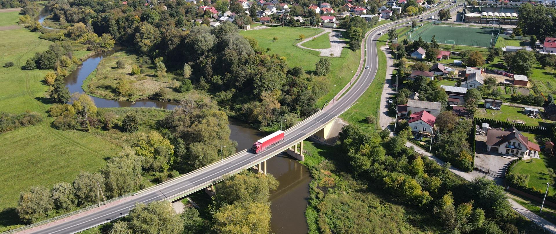 Widok z lotu ptaka na most na rzece. Zielone brzegi z bujną roślinnością. Po moście jedzie ciężarówka. W tle widoczne zabudowania mieszkalne.