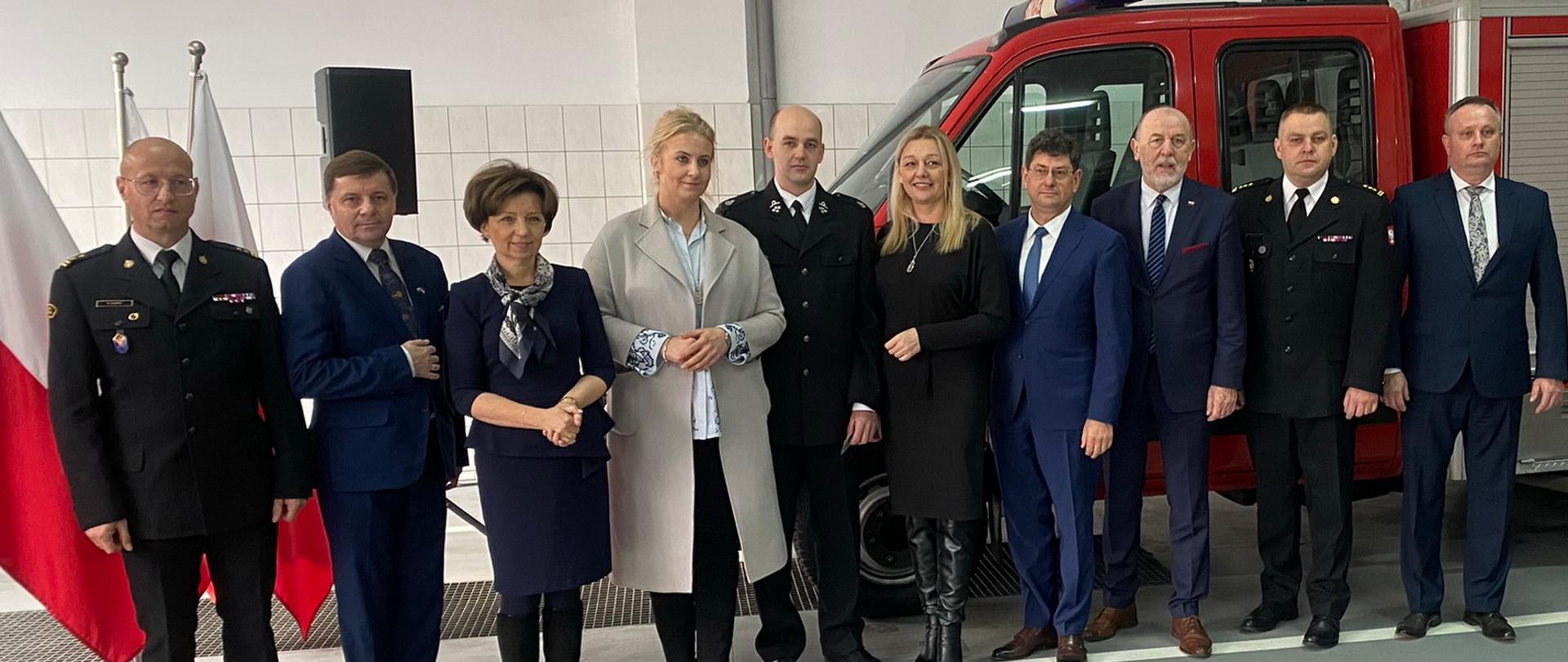 zdjęcie grupowe strażaków, kobiet i mężczyn w pomieszczeniu - garaż, w tle pojazd pożarniczy oraz flagi biało czerwone