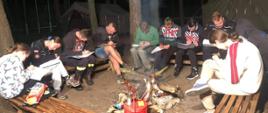Zdjęcie przedstawia grupę młodych ludzi przy ognisku w lesie, którzy piszą test w notatnikach na kolanach.