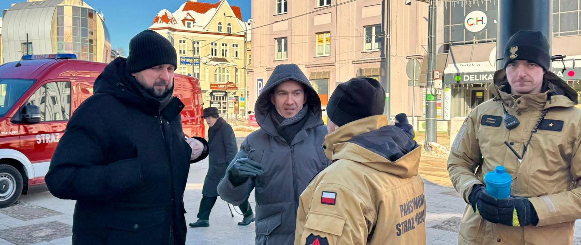 Wicewojewoda z przedstawicielami służb w Olsztynie, na zdjęciu pięciu mężczyzn, dwóch w mundurach straży pożarnej, w tle widać budynki i samochód strażacki.