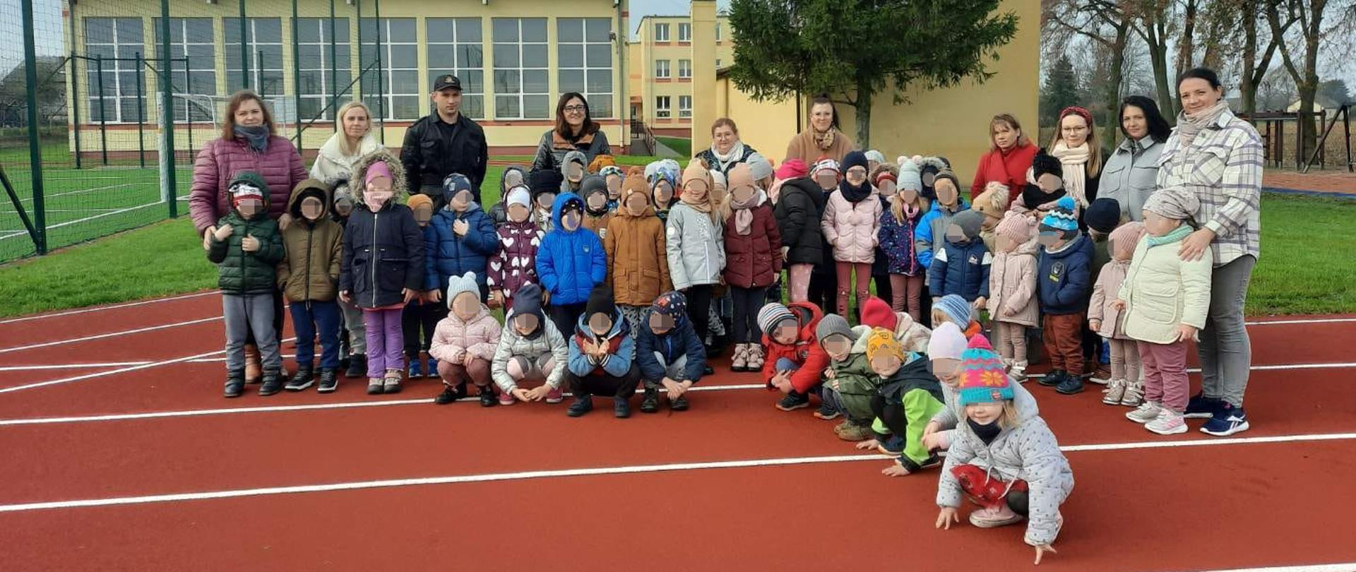 Na zdjęciu znajduje się funkcjonariusz Komendy Powiatowej Państwowej Straży Pożarnej w Rypinie, wraz z dziećmi oraz ich opiekunkami tuż po zakończonym spotkaniu dotyczącym akcji prewencyjnej - "Czujka na straży Twojego bezpieczeństwa". Są oni na bieżni boiska szkolnego, w tle znajduje się budynek szkoły.