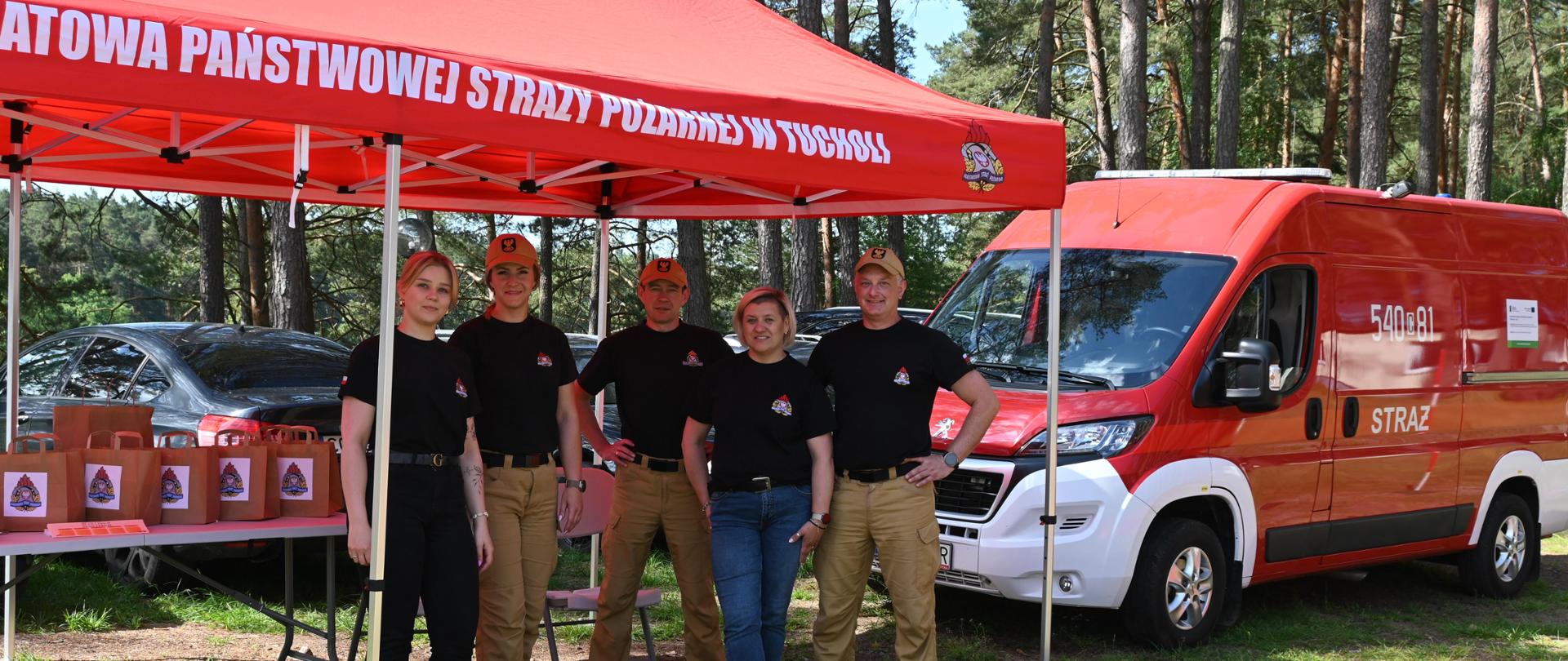 1. Na zdjęciu widać pięć osób. Od lewej strony stoi kobieta, kobieta funkcjonariusz PSP, funkcjonariusz PSP, kobieta i funkcjonariusz PSP. Strażacy mają na sobie mundur, czarne koszulki, żółte czapki z daszkiem i żółte spodnie. Wszyscy mają ubrane czarne koszulki z logo PSP. Osoby stoją pod czerwonym namiotem. Na namiocie widać biały napis ,,Komenda Powiatowa Państwowej Straży Pożarnej w Tucholi”. Namiot stoi na metalowych słupkach. Za ludźmi widać biały stolik. Na stoliku stoją brązowe torebki z logo PSP. Po prawej stronie widać czerwono-biały samochód PSP. W tle widać zaparkowane samochody oraz drzewa. Wszyscy stoją na trawie. 