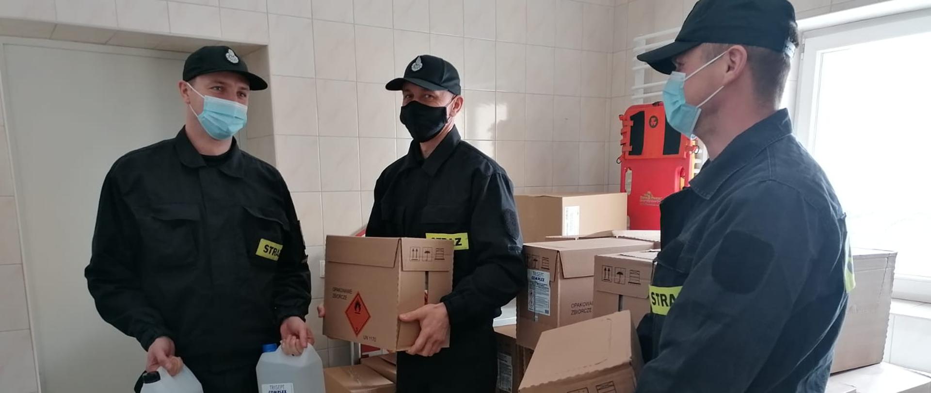 Na zdjęciu widać trzech strażaków ochotniczej straży pożarnej, którzy zostali wyposażeni w środki ochrony indywidualnej na potrzeby działań związanych z Covid-19. Zdjęcie wykonane w porze dziennej w pomieszczeniu socjalnym.