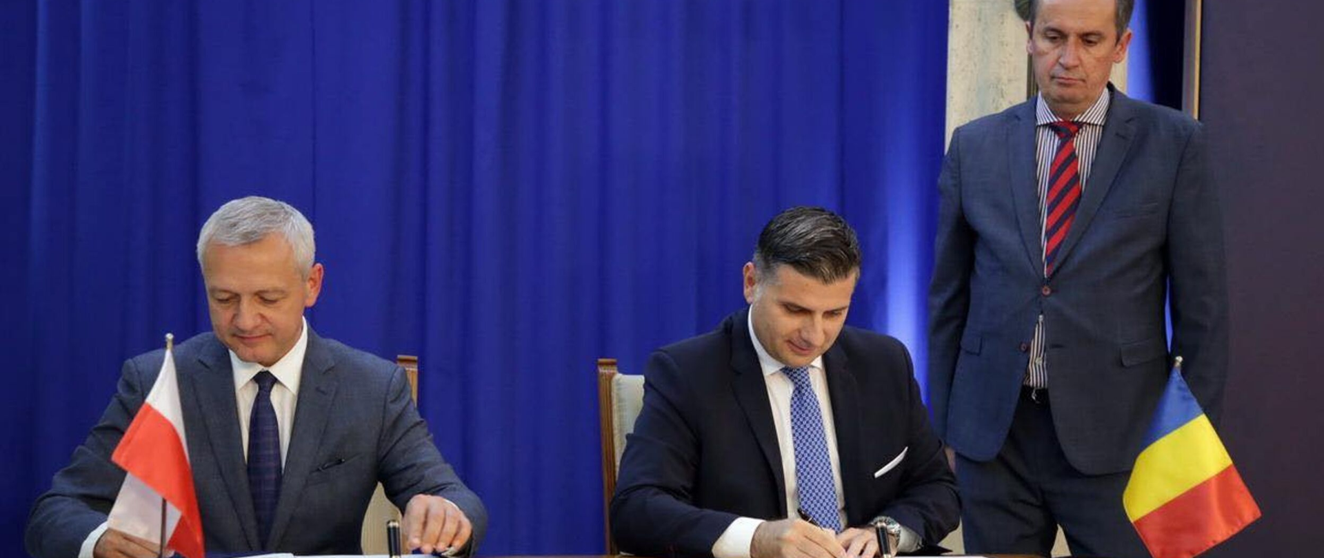 Dwóch ministrów siedzi przy stole i podpisuje dokumenty. Na blacie stoją mini flagi Polski i Rumunii. Obok stołu stoi inny mężczyzna