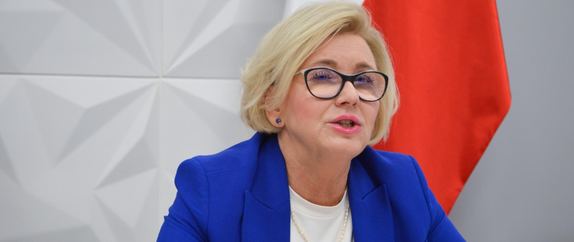 Wiceminister Machałek siedzi przy białym biurku i mówi, za nią biała ściana i polska flaga.