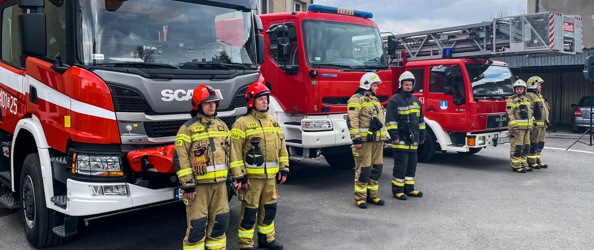 Zdjęcie przedstawia plac komendy na którym ustawione są dwa samochody ratowniczo- gaśnicze oraz drabina mechaniczna, a przed nimi stoją strażacy PSP oraz OSP