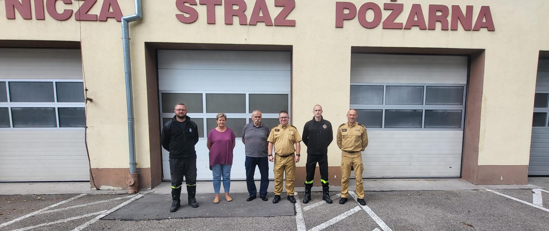 Zastępca Pomorskiego Komendanta Wojewódzkiego stoi z dwoma strażakami z OSP Pelplin oraz prezesem, przedstawicielką gminy oraz Zastępcą Komendanta Powiatowego PSP w Tczewie, przed garażami. W tle białe bramy i napis Ochotnicza Straż Pożarna.