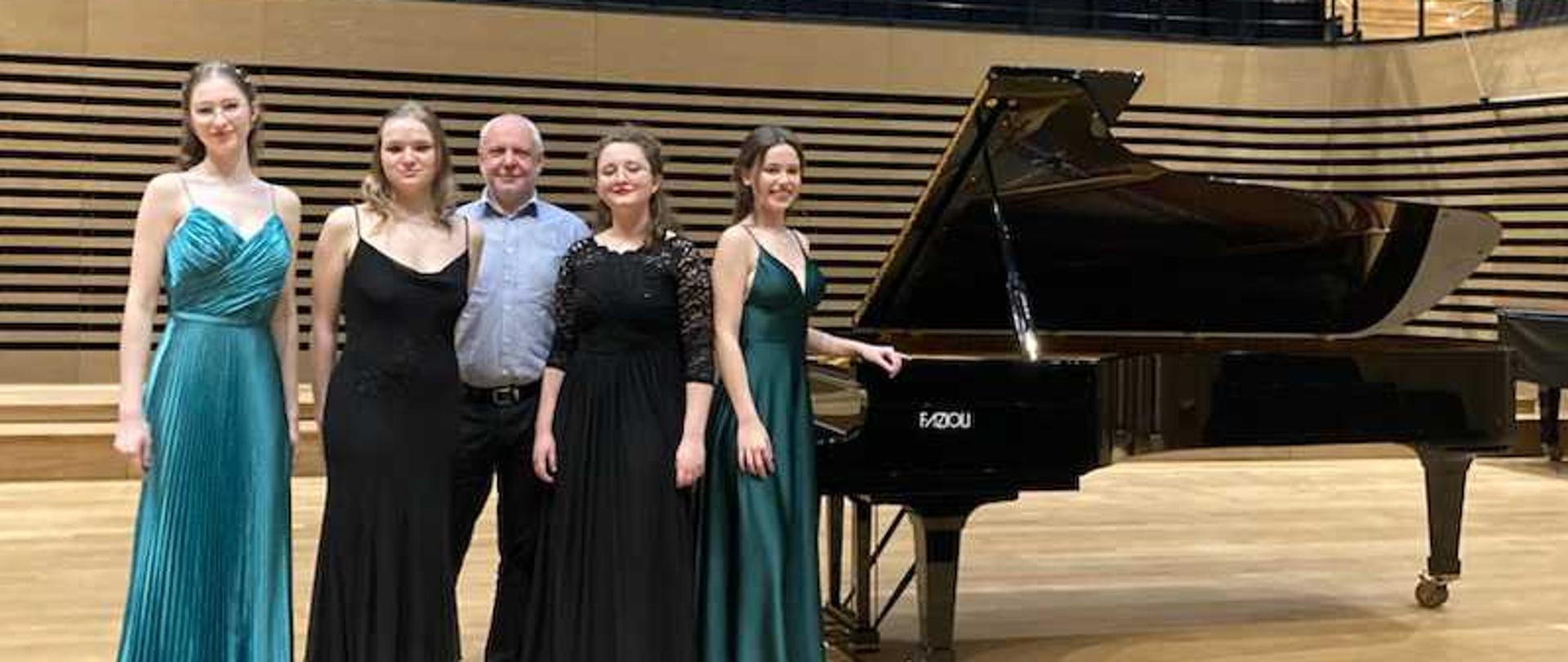 Cztery młode kobiety w długich eleganckich sukniach i mężczyzna stoją przy fortepianie Fazioli na scenie sali koncertowej PSM w Jastrzębiu-Zdroju