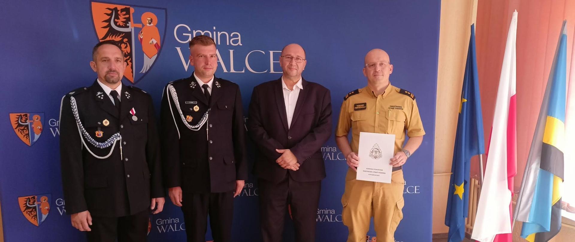 Na tle ścianki - bannera promocyjnego Gminy Walce w kolorze niebieskim z herbem Walec ustawieni sygnatariusze porozumienia: od lewej naczelnik OSP Walce, prezes OSP Walce, wójt Walec i Komendant Powiatowy PSP w Krapkowicach. Strażacy OSP w mundurach galowych, wójt Walec w ciemnym garniturze, Komendant Powiatowy PSP w umundurowaniu służbowym koloru piaskowego. Po prawej stojak z flagami : Polski, Gminy Walce i UE. 
