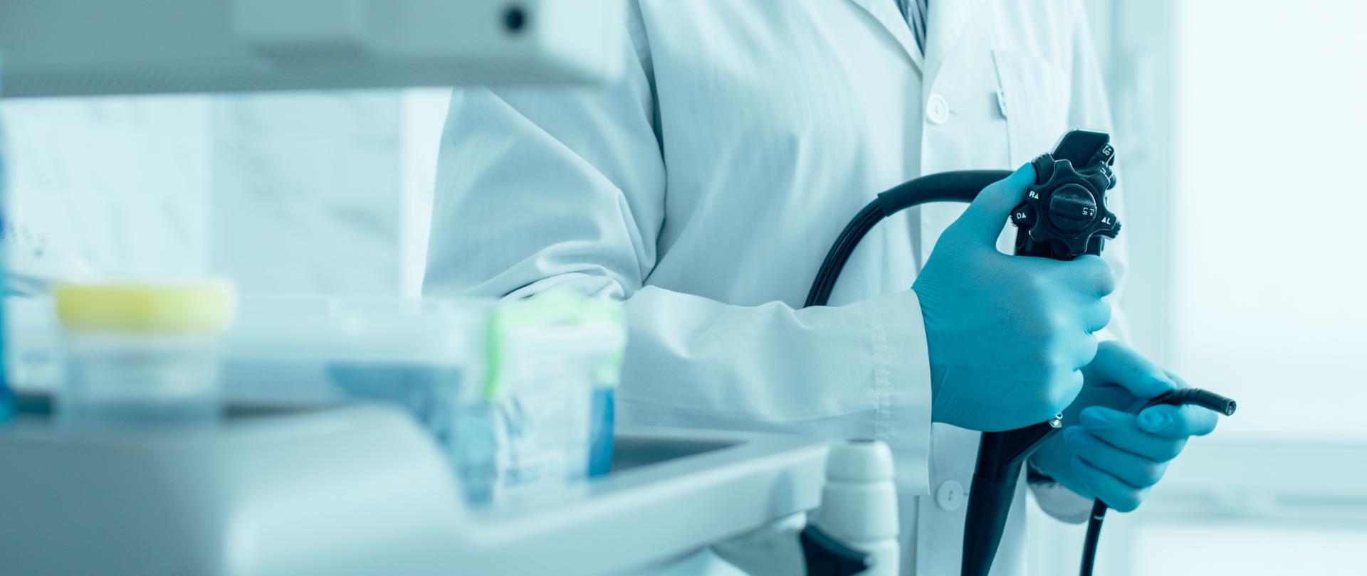 postać lekarza widziana częściowo, trzymającego narzędzie diagnostyczne do kolonoskopii