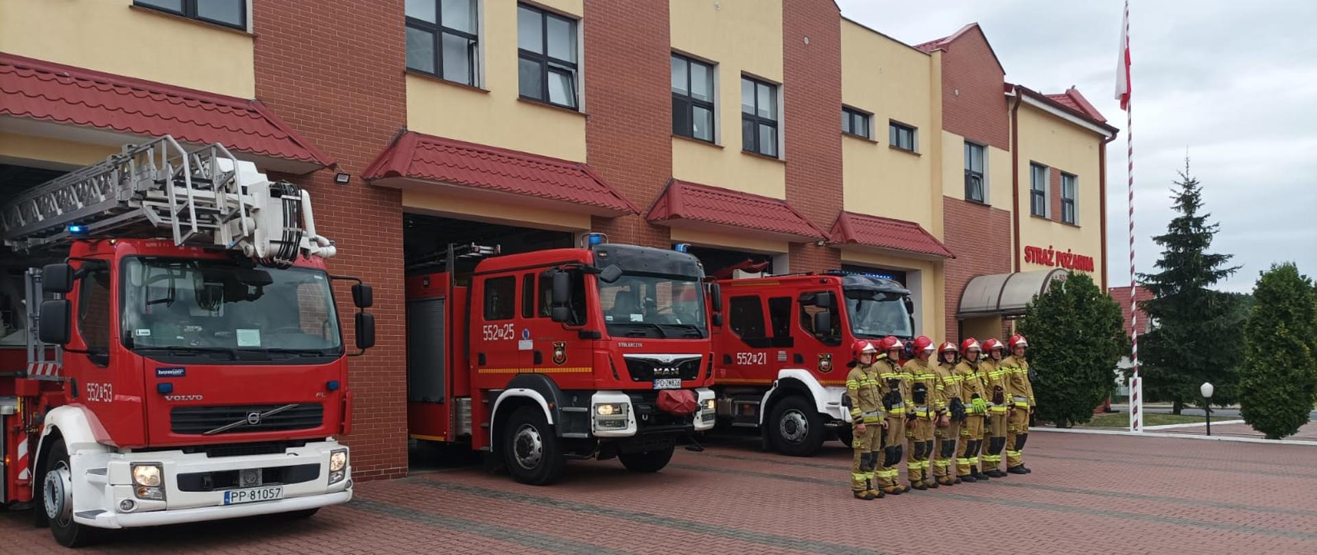Zdjęcie przedstawia strażaków stojących w szeregu na tle budynku komendy i samochodów pożarniczych.