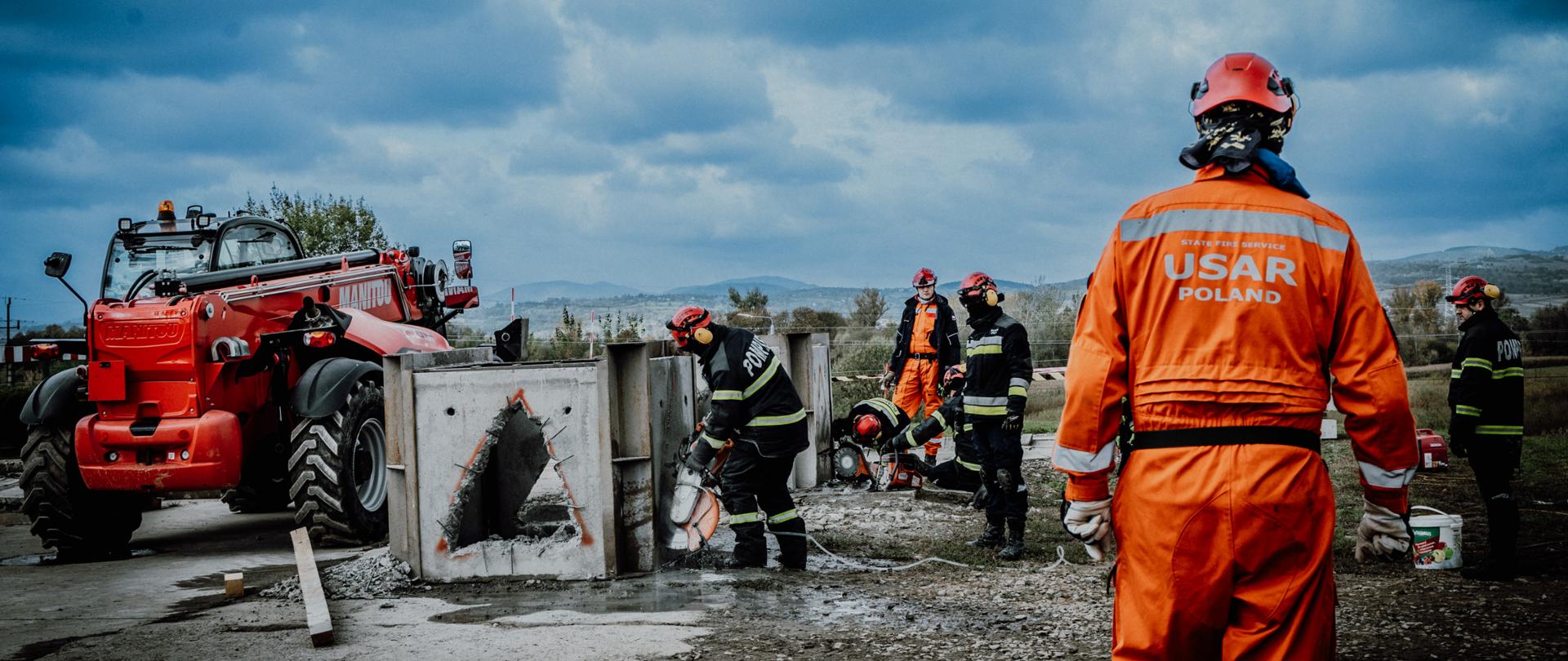 Ratownicy prowadzą ćwiczenia na stanowisku z betonowymi elementami konstrukcyjnymi. Na pierwszym planie stoi członek USAR Poland w pomarańczowym kombinezonie, obserwujący działania zespołu.