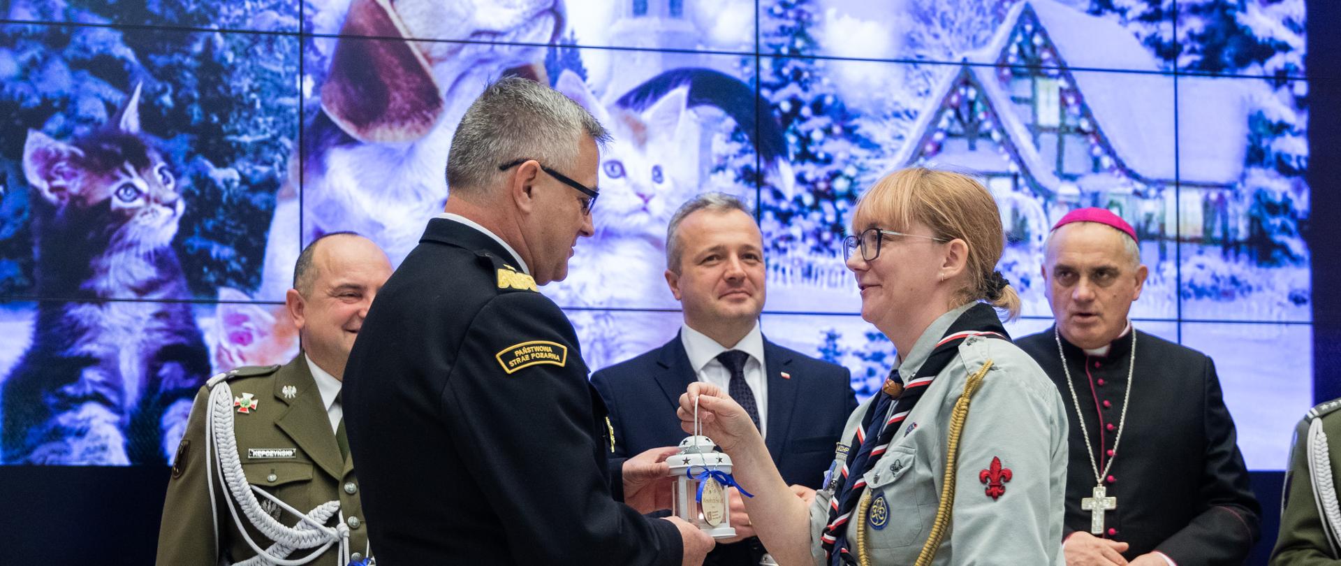 Na pierwszym planie komendant wojewódzki i druhna harcmistrz. Przekazują sobie symboliczne światełko pokoju. W tle wojewoda, biskup, żołnierz...