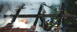 Na zdjęciu widać spaloną konstrukcję więźby dachowej i dwóch strażaków w aparatach ochrony dróg oddechowych pracujących na poddaszu.
