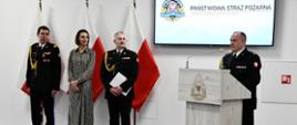 Strażak w mundurze wyjściowym stoi i przemawia przy mównicy za nim stoi kadra kierownicza Państwowej Straży Pożarnej wojewoda pomorska ustawione są flagi Polski na ścianie wisi rota ślubowania ekran z logiem i napisem Państwowej Straży Pożarnej oraz zegar.