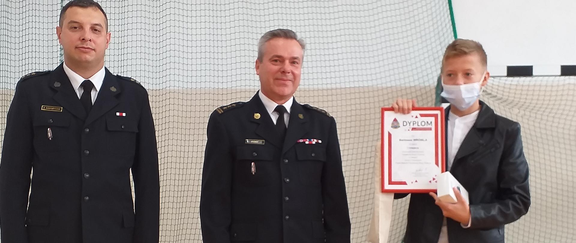 Zdjęcie przedstawia dwóch funkcjonariuszy w mundurach wyjściowych z uczniem, który wygrał konkurs kalendarzowy, uczeń prezentuje nagrodę rzeczową i dyplom, w tle bramka sportowa w sali gimnastycznej