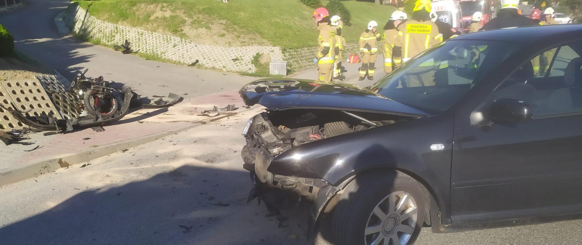 Zdjęcie przedstawia przednią część samochodu osobowego w kolorze czarnym, który w wyniku zderzenia z motocyklem posiada rozbity przód. Z lewej strony widoczny jest zniszczony motocykl, elementy z pojazdów leżące na jezdni i na chodniku. W prawej górnej części znajdują się strażacy w umundurowaniu specjalnym, częściowo widoczna karetka pogotowia ratunkowego i samochód strażacki.