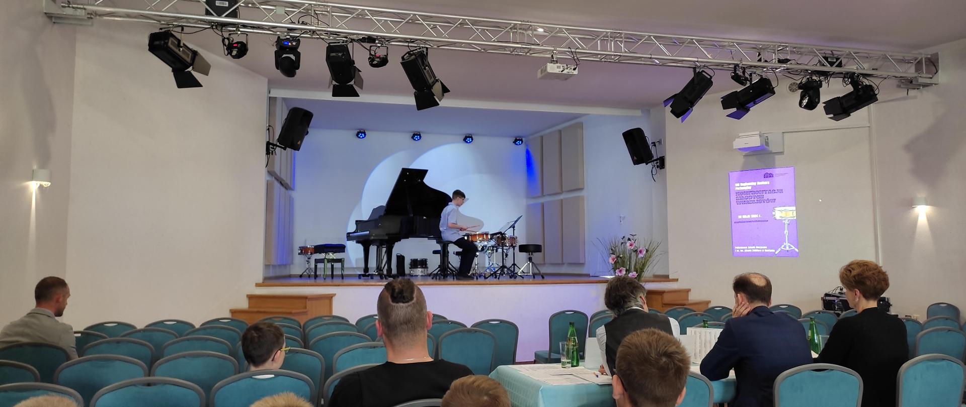 Uczestnicy konkursu oraz jury siedzący na widowni. Na scenie uczeń podczas swojej konkursowej prezentacji w grze na werblu. 