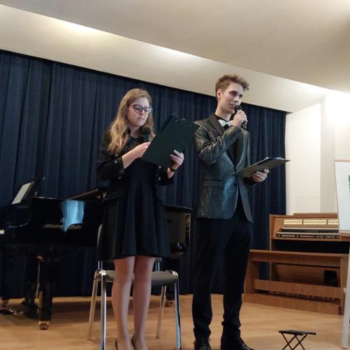 Dwoje uczniów stoi na scenie i zapowiada koncert. W tle fortepian i elektroniczne organy. 