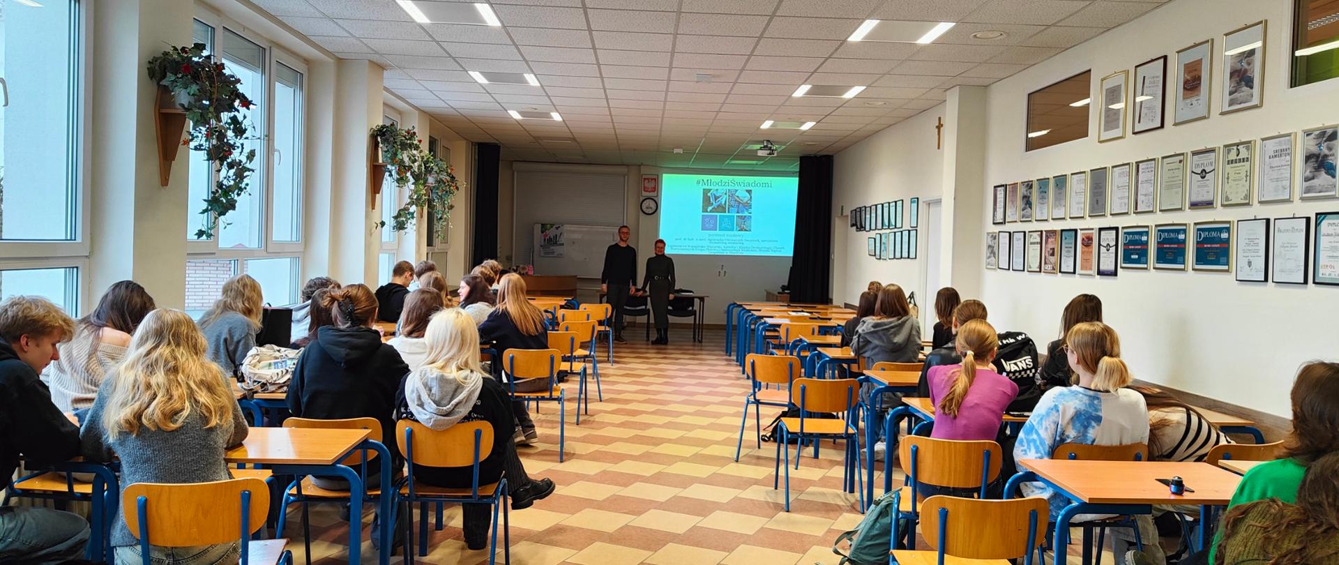 Edukacja młodzieży w szkole w ramach bezpiecznych ferii. Uczniowie siedzą w ławkach, słuchają prelegentów i oglądają prezentację multimedialną. 