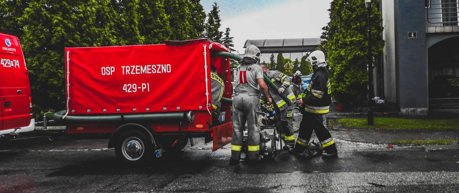 Strażacy w jasnych i ciemnych ubraniach ładują do czerwonej przyczepy strażackiej pompę. W tle zielone tuje i szary budynek.