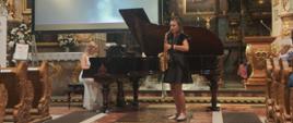 Młoda kobieta w czarnej sukience gra na saksofonie podczas koncertu w kościele. Za nią, przy fortepianie, siedzi akompaniatorka w białej sukni. W tle widać ołtarz i ekran projekcyjny.