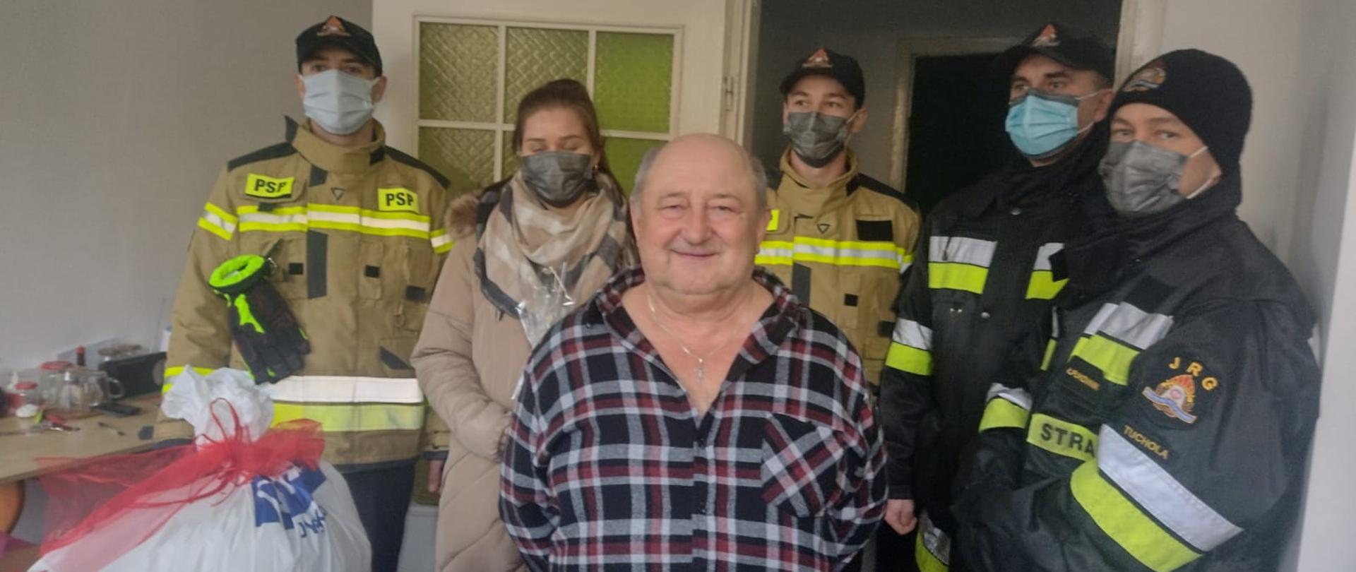 Zdjęcie przedstawia 4 strażaków i pracownika cywilnego KPPSP w Tucholi w mieszkaniu, do którego dostarczono prezenty w ramach akcji „Świąteczna Tucholska Paczka” organizowanej przez Miejski Ośrodek Pomocy Społecznej w Tucholi. Na pierwszym planie zdjęcia widać Pana Krzysztofa i prezenty, które otrzymał. 