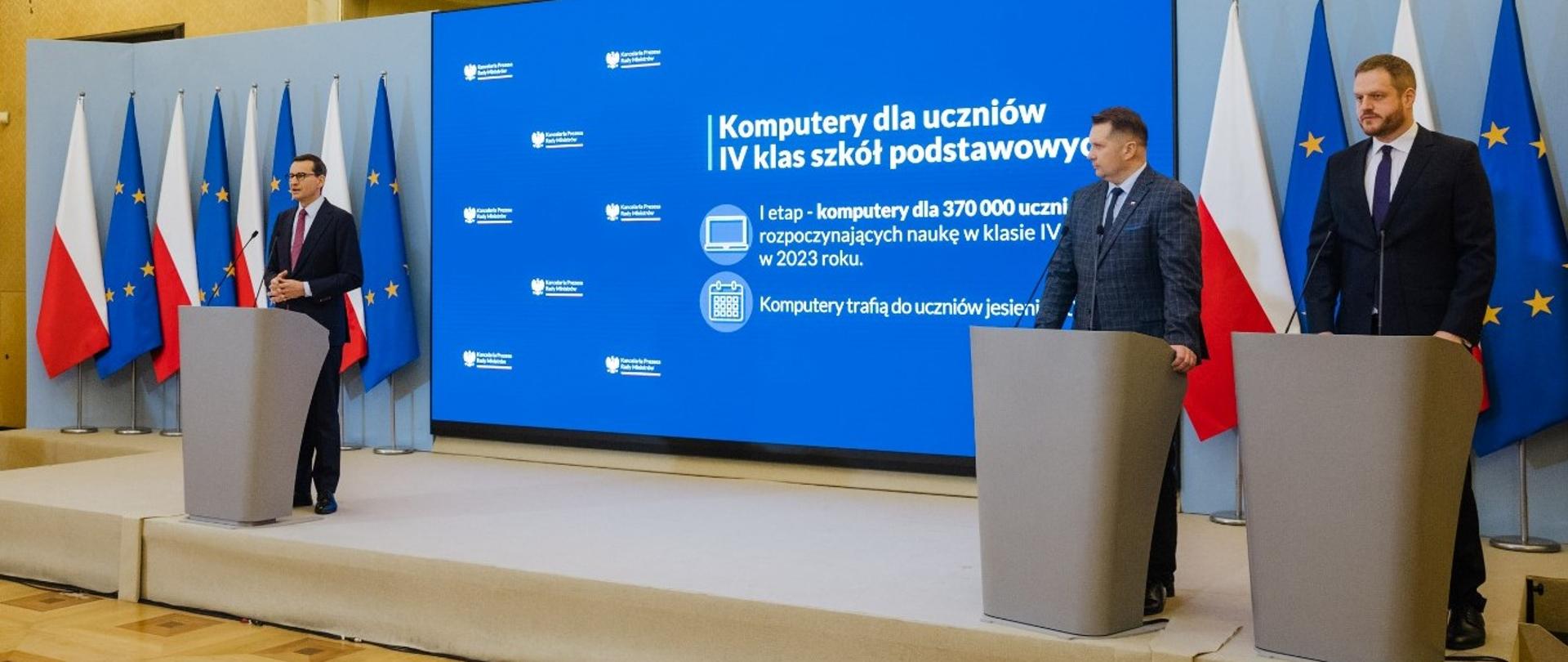 Na podwyższeniu przy szarych mównicach stoi trzech mężczyzn w garniturach, za nimi niebieska ściana z napisem Komputery dla uczniów IV klas szkół podstawowych.