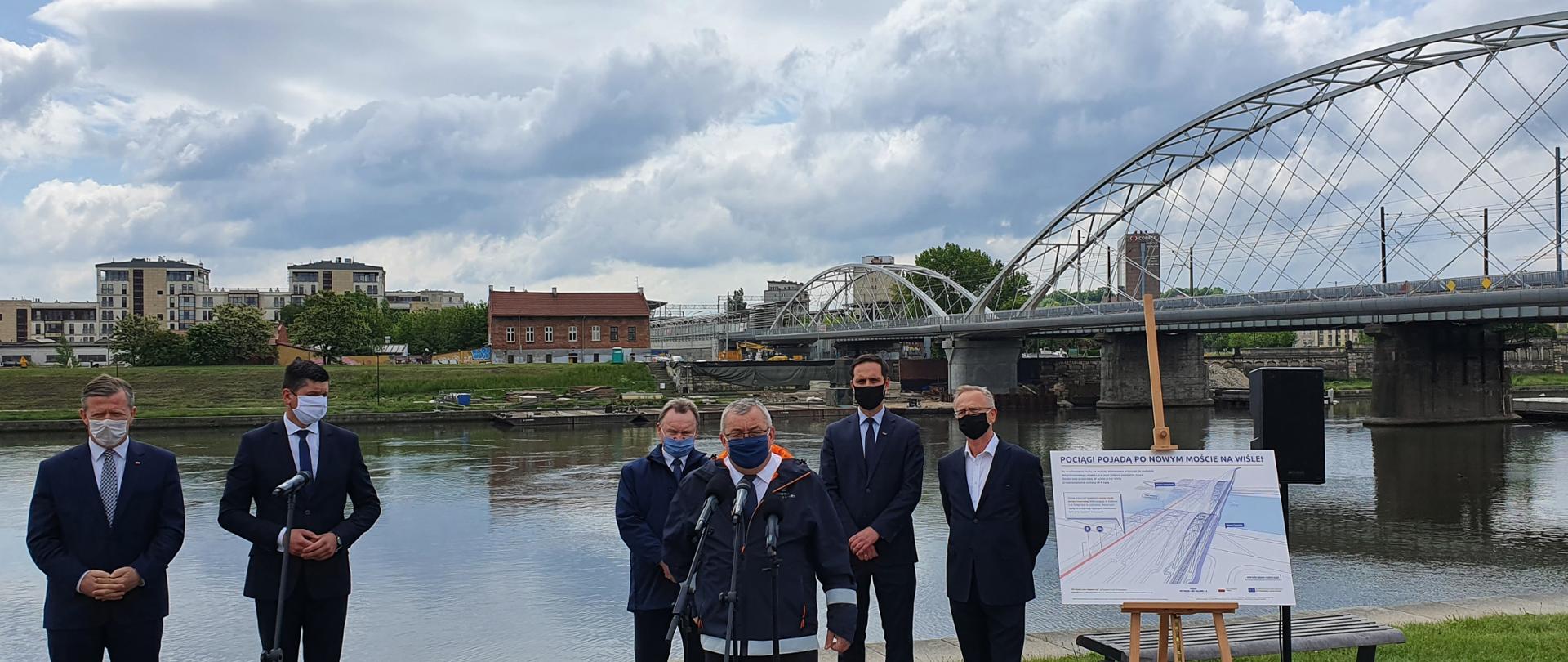 Na zdjęciu widać 6 mężczyzn, którzy stoją przy bulwarach nad rzeką w Krakowie. W tle widać nowy most kolejowy. Pogoda jest słoneczna, niebo błękitne.