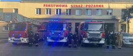 Na zdjęciu widoczni strażacy przy wozach bojowych koloru czerwonego. W samochodach uruchomione są sygnały świetlne. Strażacy stoją równo z samochodami w umundurowaniu koszarowym oraz służbowym na baczność. W tle widoczny budynek JRG nr 1 w Świnoujściu. 