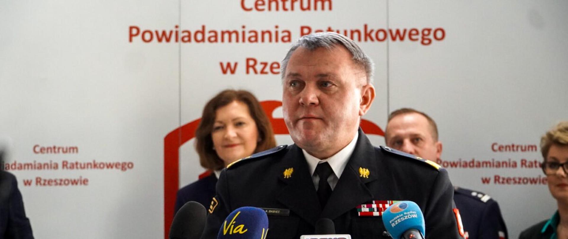 Zdjęcie zrobione w pomieszczeniu Centrum Powiadamiania Ratunkowego w Rzeszowie podczas konferencji prasowej. Do mikrofonu przemawia podkarpacki komendant wojewódzki PSP. Z tyłu stoją uczestnicy konferencji.