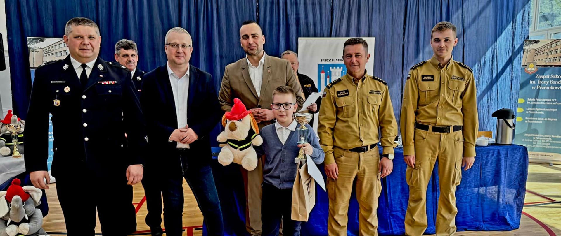 Zdjęcie przedstawia oficjalną uroczystość wręczenia nagród, która odbywa się na sali gimnastycznej (widoczny jest parkiet z liniami boiska). W centrum uwagi znajduje się młody chłopiec trzymający puchar, dyplom oraz torbę z upominkiem, otoczony przez grupę dorosłych mężczyzn. Oto szczegółowy opis osób i otoczenia na zdjęciu: Osoby na zdjęciu Chłopiec w centrum: Nosi okulary, ciemne spodnie, jasną koszulę z kołnierzykiem i szary sweter lub kamizelkę. Trzyma w dłoniach trofeum (puchar) oraz papierową torbę podarunkową. Mężczyźni w mundurach strażackich (po lewej stronie): * Na pierwszym planie stoi mężczyzna w ciemnogranatowym lub czarnym mundurze wyjściowym z przypiętymi odznaczeniami. Za nim, nieco w głębi, widać kolejnego mężczyznę w podobnym ciemnym umundurowaniu. Mężczyźni w strojach cywilnych (w środku): Po lewej stronie chłopca stoi mężczyzna w okularach, ciemnej marynarce, jasnej koszuli i jeansach. Tuż za chłopcem stoi mężczyzna w jasnobrązowej (beżowej) marynarce i białej koszuli, który trzyma pluszową maskotkę psa w strażackim hełmie. Mężczyźni w piaskowych mundurach (po prawej stronie): Obok chłopca stoi mężczyzna w oficjalnym, piaskowym (beżowym) mundurze strażackim (charakterystycznym dla Państwowej Straży Pożarnej). Po jego prawej stronie stoi młodszy mężczyzna w identycznym piaskowym umundurowaniu. Tło i otoczenie Sceneria: W tle zawieszona jest duża, niebieska lub granatowa kotara, która zasłania ścianę sali. Materiały promocyjne: * Po prawej stronie widoczny jest pionowy baner (roll-up) z logo, zdjęciem budynku oraz napisami w języku polskim (można odczytać fragmenty wskazujące na szkołę lub zespół szkół im. Ireny Sendlerowej). W tle widać również białą tablicę lub plakat z herbem miasta (widoczna baszta) i napisem zaczynającym się od "PRZEM...". Elementy dodatkowe: Po lewej stronie na podłodze leży kolejna, większa pluszowa maskotka (słoń w strażackim hełmie) oraz widoczne są inne nagrody lub puchary na stoliku pokrytym obrusem. Cała scena ma charakter oficjalnego, uroczystego rozstrzygnięcia konkursu lub turnieju – najprawdopodobniej o tematyce strażackiej lub związanej z bezpieczeństwem.