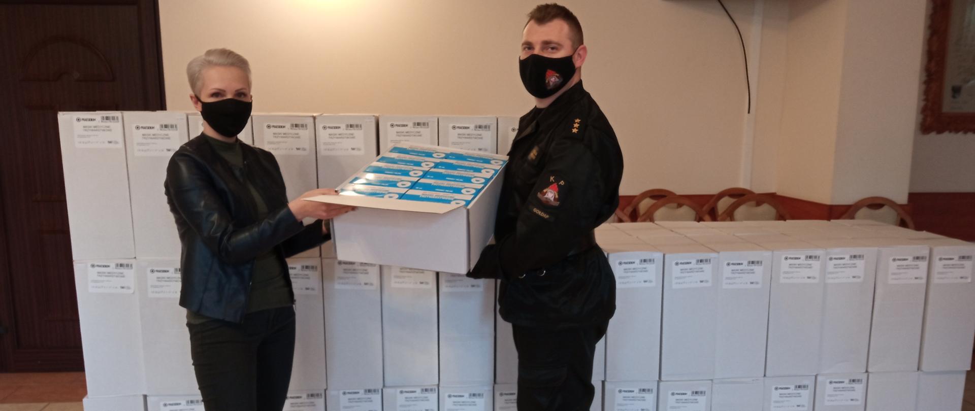 Zdjęcie przedstawia symboliczne przekazanie przez p.o. Zastępcę Komendanta Powiatowego PSP w Gołdapi kpt. Michała Herman kartonu z 1000 szt. maseczek ochronnych dla Kierownik wydziału ds. administracyjnych UM w Gołdapi Pani Róży Popławskiej