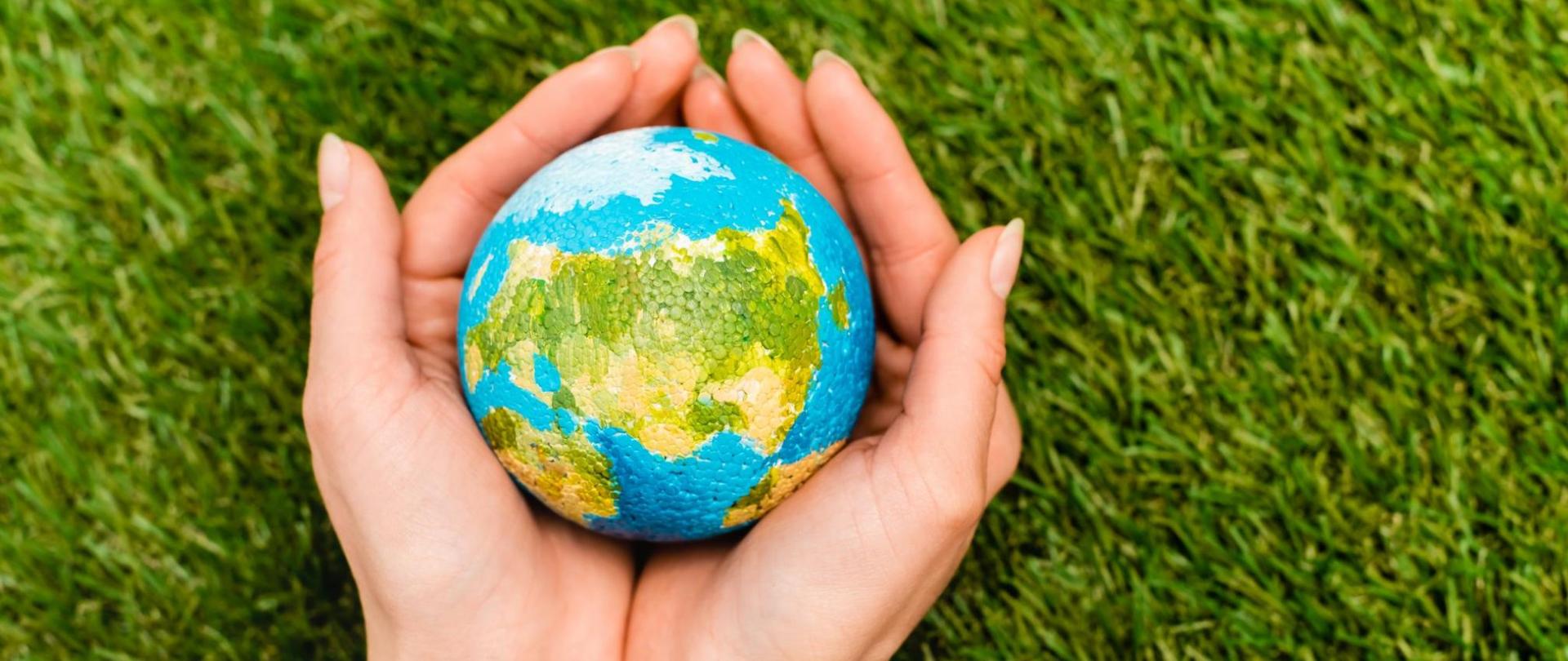 Na zdjęciu są dłonie kobiety a w nich miniatura planety Ziemia. W tle jest trawa.