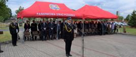W dwóch rzędach pod czerwonymi namiotami z napisem Państwowa Straż Pożarna stoją zaproszeni goście, a przed nimi stoi Komendant Wojewódzki przemawiający przez mikrofon.