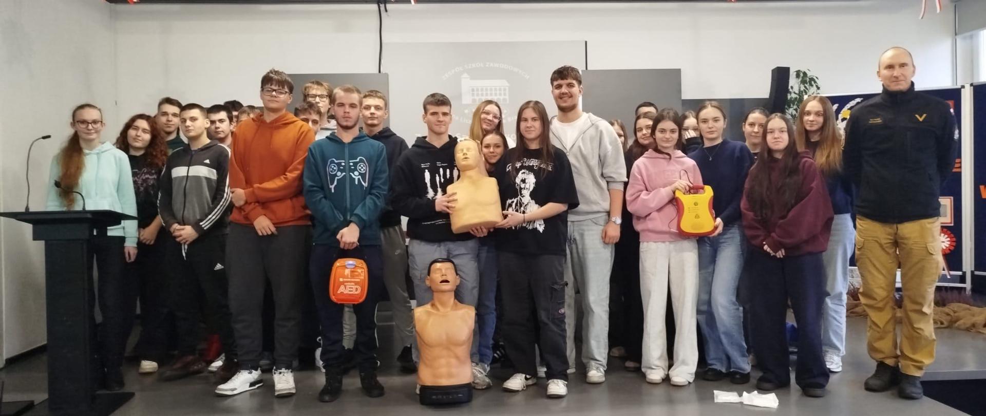 Zdjęcie przedstawia grupę młodzieży wraz z instruktorem podczas szkolenia z zakresu pierwszej pomocy. Uczestnicy stoją na scenie, a przed nimi umieszczone są fantomy do nauki resuscytacji krążeniowo-oddechowej oraz defibrylatory AED. W tle widać dekoracje w barwach narodowych, co może wskazywać, że wydarzenie odbyło się w okolicach święta narodowego. Atmosfera jest edukacyjna i pozytywna – uczestnicy wydają się zaangażowani i zainteresowani tematem.