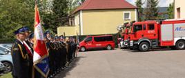 Zdjęcie przedstawia poczet sztandarowy oraz strażaków, którzy stoją w dwuszeregu na placu Komendy Powiatowej PSP w Człuchowie. W tle pojazdy pożarnicze.