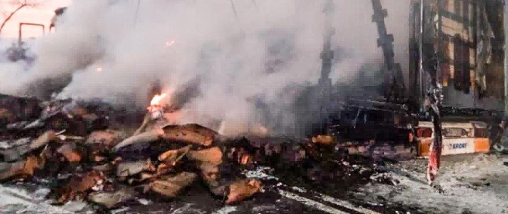 Zdjęcie przedstawia pożar ciągnika siodłowego z naczepą wypełnioną opakowaniami tekturowymi, pożar w fazie rozwiniętej, ładunek rozsypał się na drogę, duże zadymienie. 