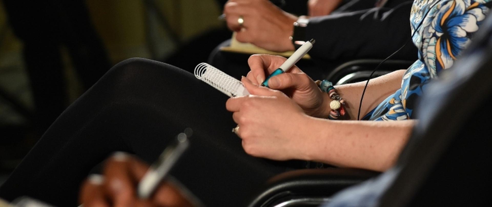 Zbliżenie na ręce osób robiących notatki podczas spotkania; widoczne długopisy, zeszyty i biżuteria.