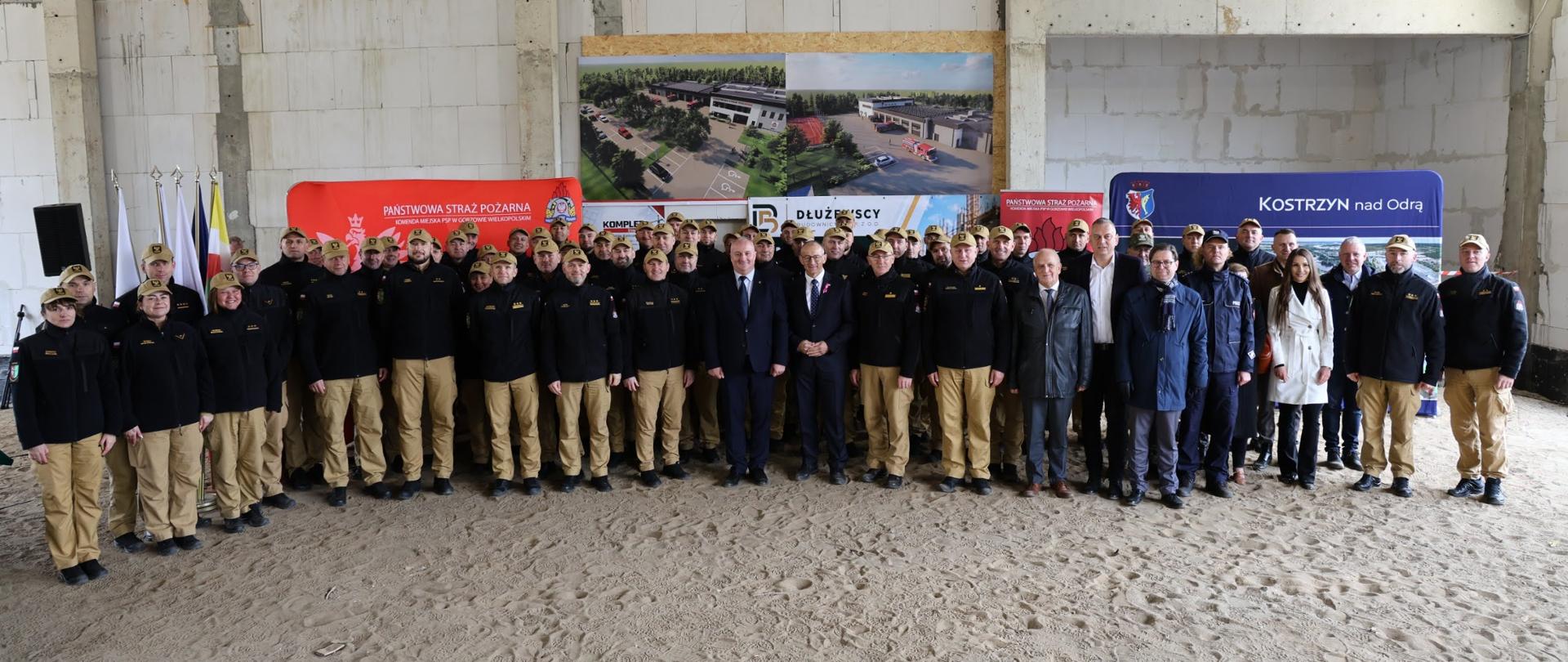 Liczna grupa strażaków i osób w ubraniach cywilnych pozuje do wspólnego zdjęcia wewnątrz budynku w stanie surowym. W tle flagi, wizualizacje strażnicy oraz banery Państwowej Straży Pożarnej i miasta Kostrzyn nad Odrą.